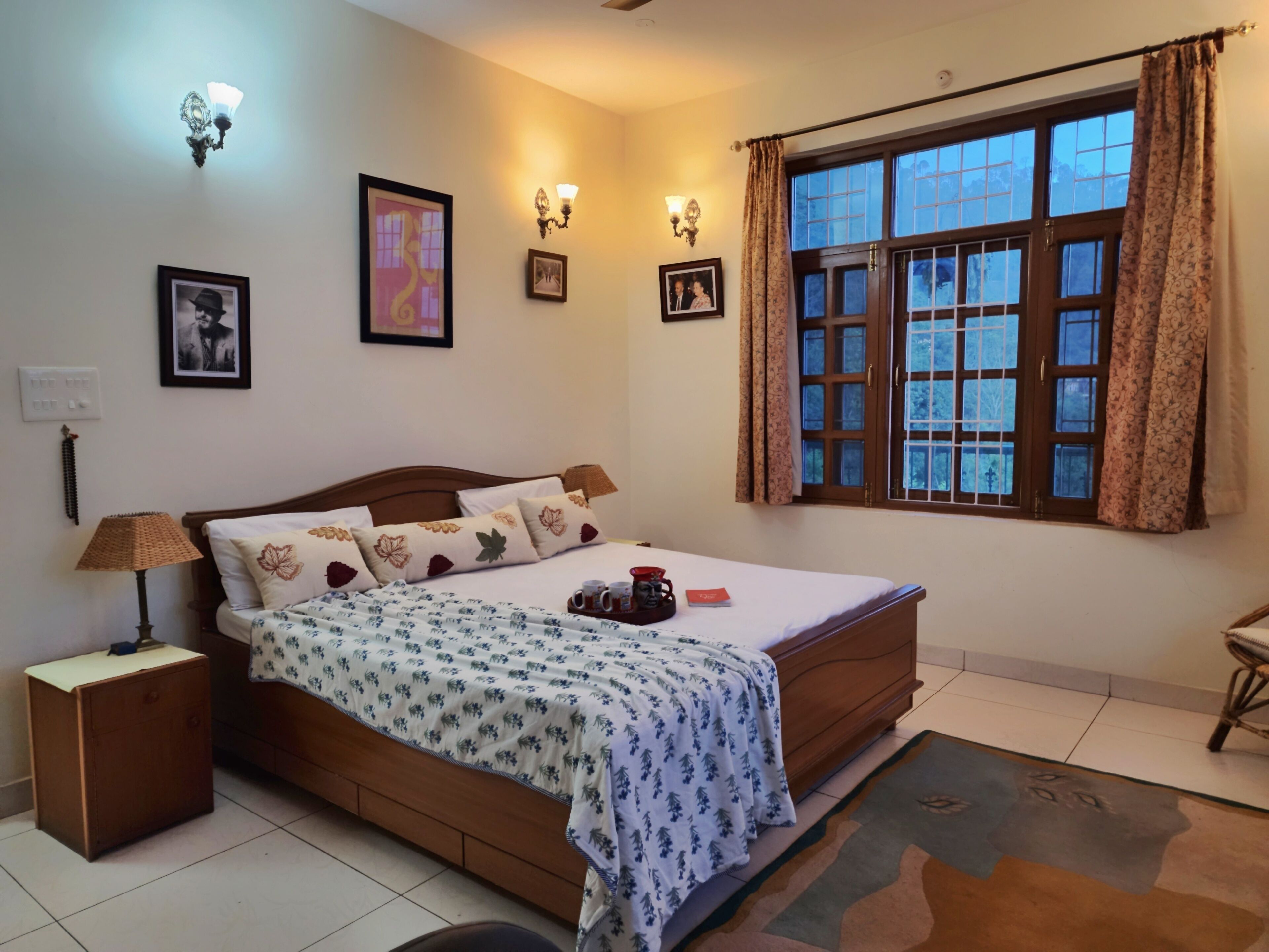 A bedroom with a wooden bed, a nightstand, a lamp, a window with curtains, and framed pictures on the wall.