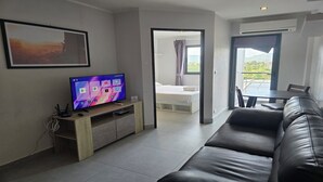 A modern living room with a flat-screen TV, a black leather sofa, a dining table, and a view of the outdoors through a large window. - The Chalong Pearl 1BR  T1-703 (Chalong)
