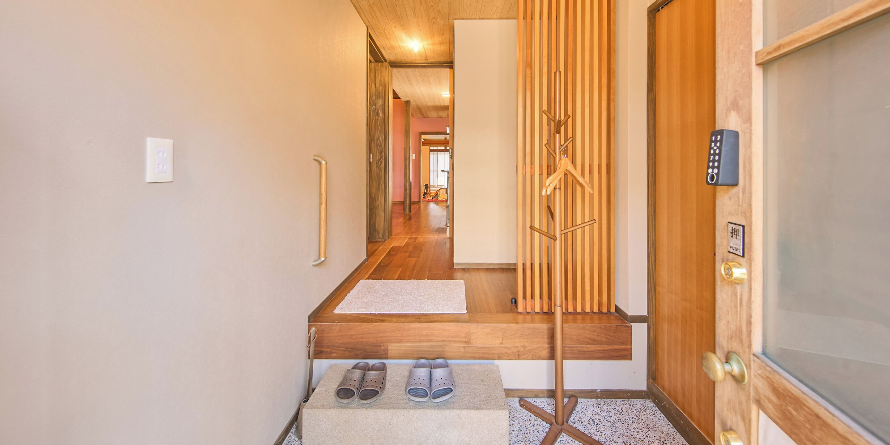 A hallway with a wooden floor, a coat rack, a door with a keypad, and a small table with shoes.