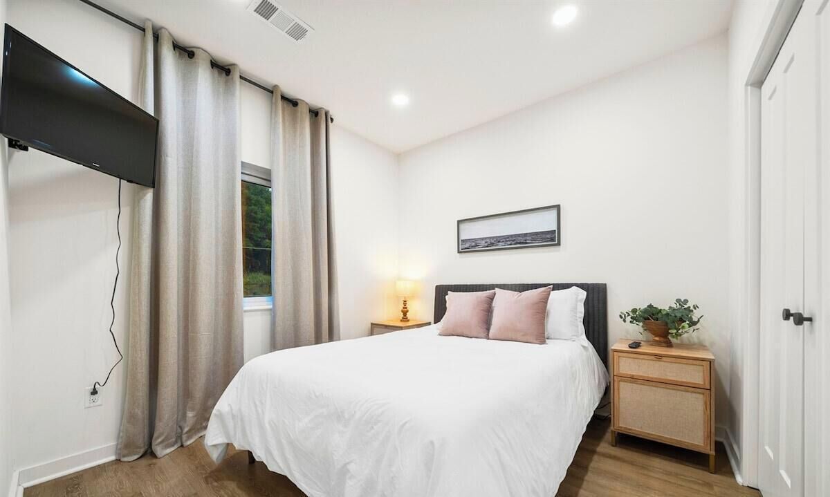 Une chambre à coucher avec un lit, une table de chevet avec une lampe, un téléviseur fixé au mur et une fenêtre avec des rideaux.