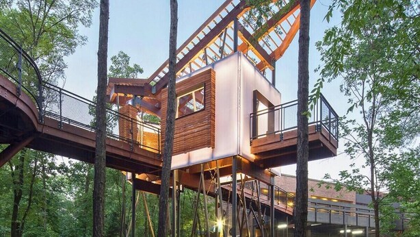 Een boomhuis met een houten buitenkant, gebouwd op poten en verbonden door een brug.