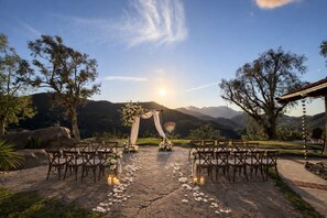 Hochzeitzeremonie im Freien mit einem Kiesweg, Holzsitzen und floralen Dekorationen vor einer Bergkulisse.