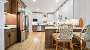 A modern kitchen with wooden cabinets, stainless steel appliances, and a central island with wicker chairs. - Side-by-Side Lake Hamilton Homes w/Dock, Pool (Hot Springs)