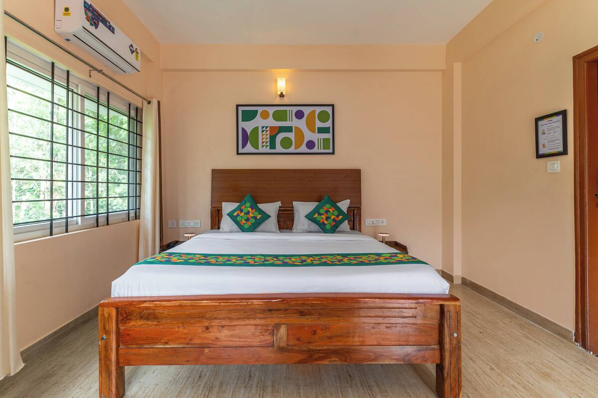 Un dormitorio con una cama de madera, un ventanal con barrotes, un aire acondicionado fijado a la pared y un cuadro abstracto enmarcado.