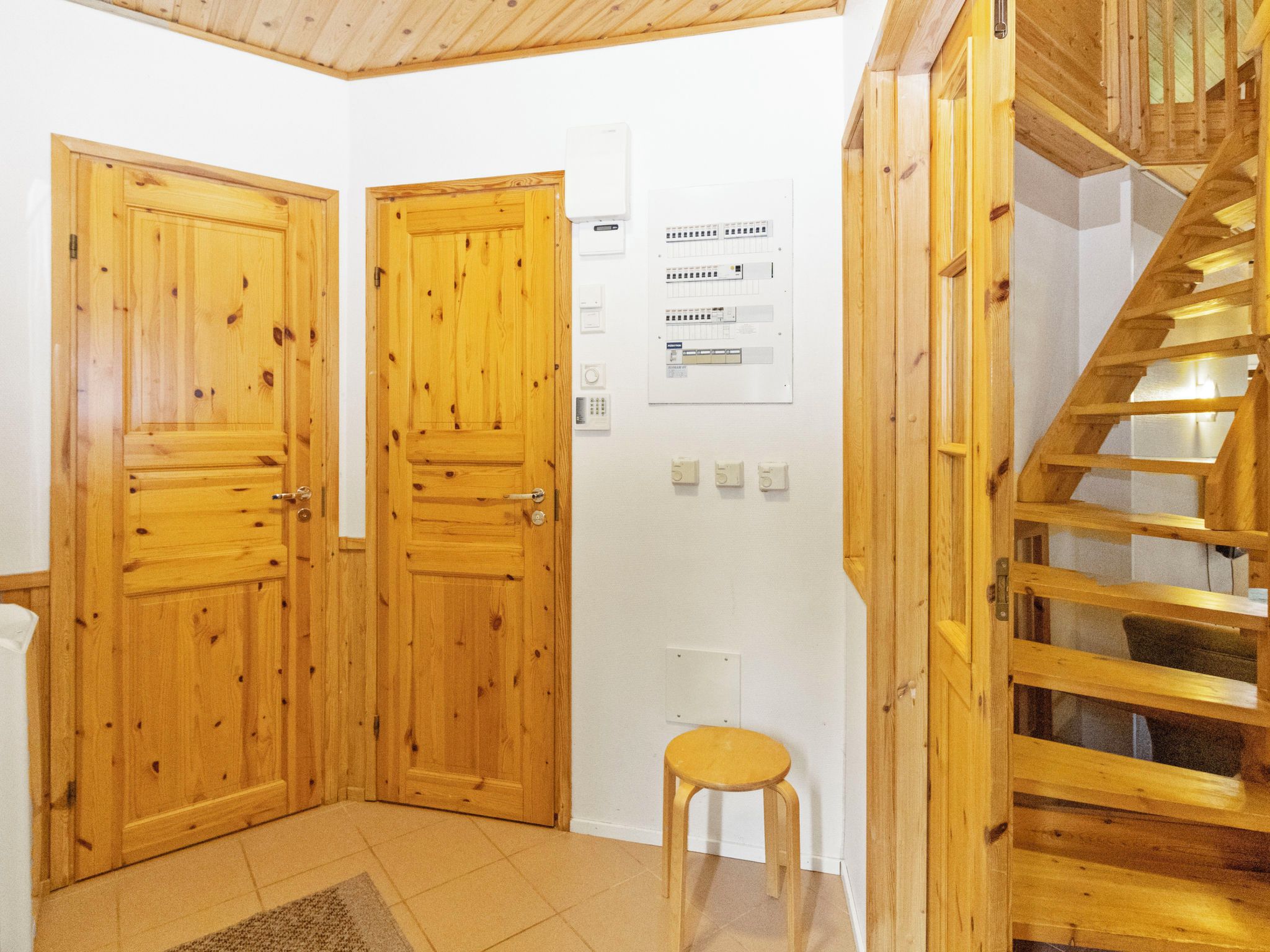 A room with wooden doors, a small stool, a wall-mounted control panel, and a wooden staircase.