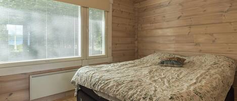 A bedroom with a bed, a window with blinds, wooden walls, and a radiator.