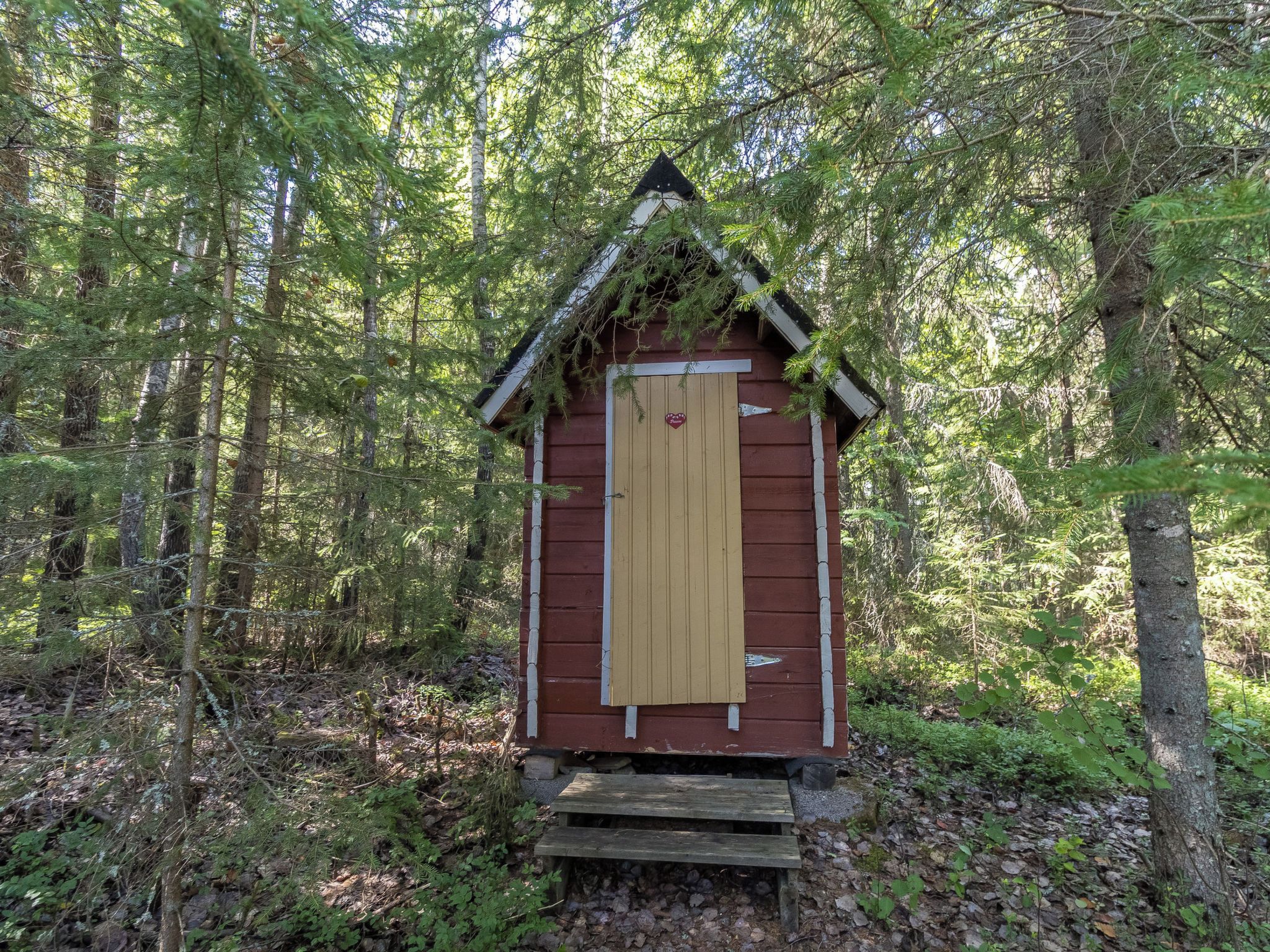 Pieni puinen rakennelma, jossa on kalteva katto, ja joka sijaitsee tiheässä metsässä, jossa on runsaasti puita ja aluskasvillisuutta.