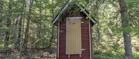 A small wooden structure with a sloped roof, situated in a forested area with dense trees and undergrowth.