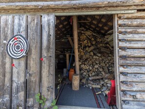 Un bâtiment rustique en bois, avec un jeu de fléchettes accroché au mur, une pile de bois de chauffage à l’intérieur et un seau en métal.
