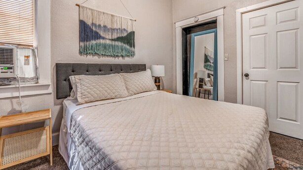 A bedroom with a bed, a wall-mounted tapestry, a bedside table with a lamp, a window with blinds, and a door. - Cozy Central Hideaway in Midtown Memphis (Memphis)