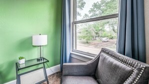Une pièce avec un mur vert, une fenêtre ornée de rideaux bleus, un fauteuil rembourré de couleur grise, une petite table avec une lampe et une plante en pot.
