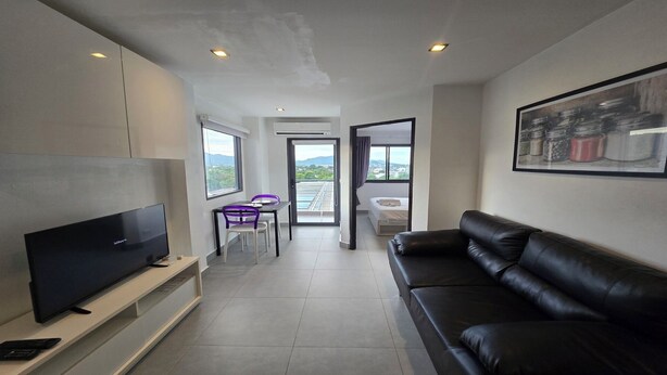 A modern living room with a flat-screen TV, a black leather sofa, and a dining area with a table and chairs. - Urban loft 1BR T1-706 (Chalong)