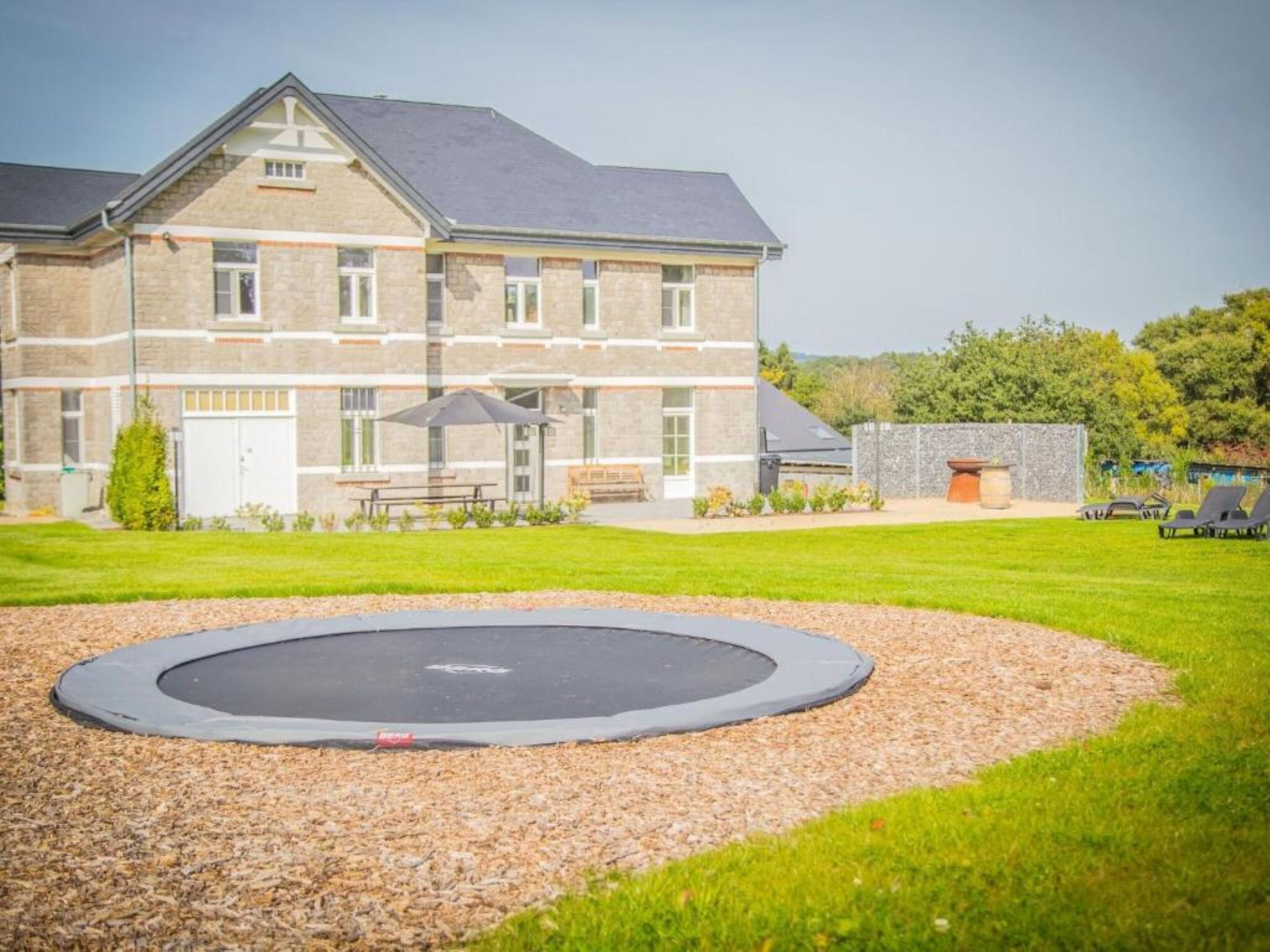 Een gebouw met een gevel van natuursteen, een gazon en een trampoline op de voorgrond.