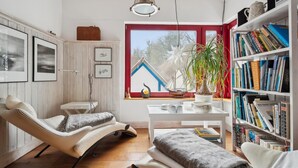 Um quarto aconchegante com teto de madeira, uma janela grande com vista para uma casa, uma estante cheia de livros e um espaço de estar confortável com uma poltrona e almofadas.