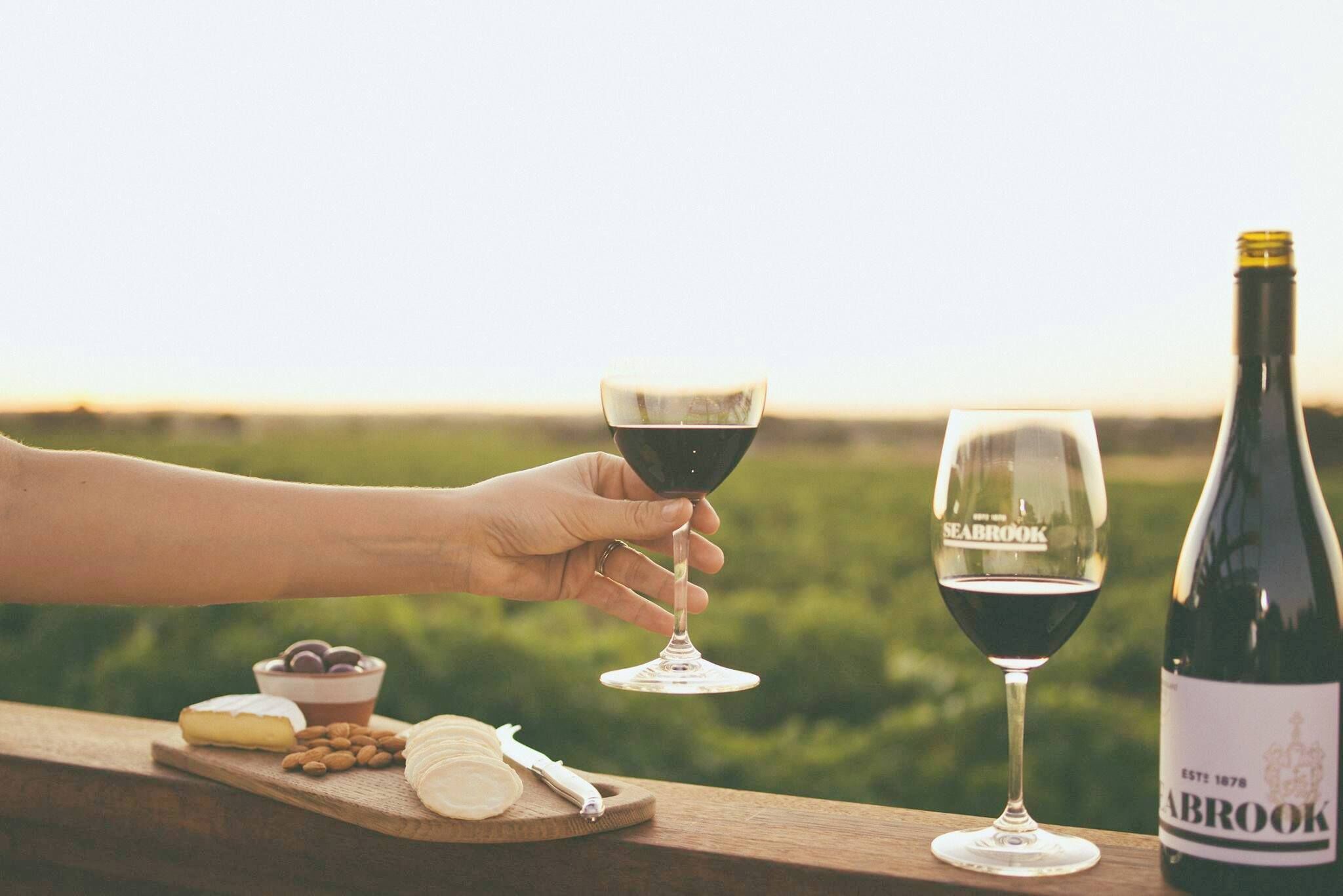 A wooden table with a bottle of Seabrook wine, two wine glasses, and a cheese board with various cheeses and nuts.