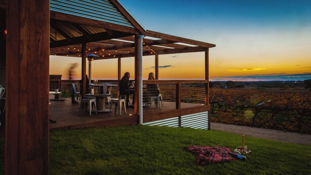 Une terrasse en bois avec des sièges extérieurs, offrant une vue sur un vignoble au coucher du soleil.