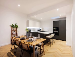 Eine moderne Küche mit einer zentralen Kochinsel, einem Essbereich mit Holztisch und -stühlen sowie einer kleinen Pflanze auf einem Regal.
