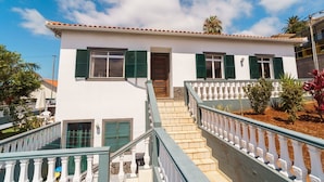 A two-story building with a white facade, green shutters, and a tiled roof. There is a staircase leading to the entrance, and a white balustrade along the railing.