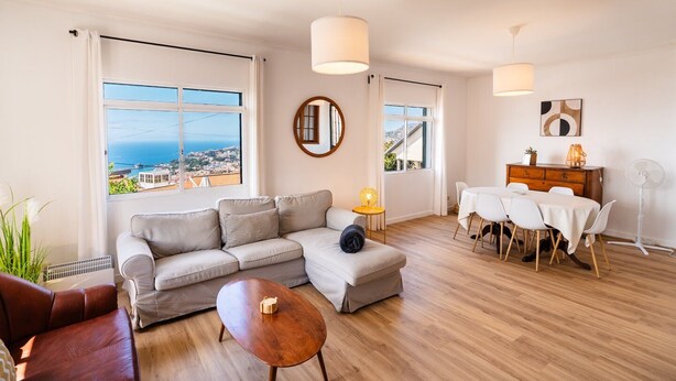 A modern living room with a sofa, a coffee table, and a dining area with a table and chairs. There are large windows offering a view of the sea.