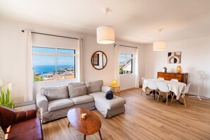 A modern living room with a sofa, a coffee table, and a dining area with a table and chairs. There are large windows offering a view of the sea. - GuestReady - Typical place in Funchal (Funchal)