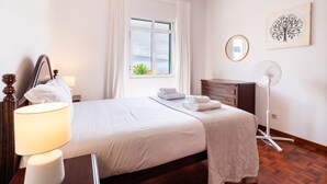 A neatly arranged hotel room with a bed, a bedside lamp, a dresser, a mirror, a window with curtains, a wall-mounted fan, and a framed artwork.