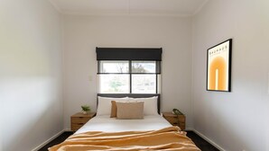 A bedroom with a bed, a window with a black valance, a framed picture on the wall, and two bedside tables with plants.