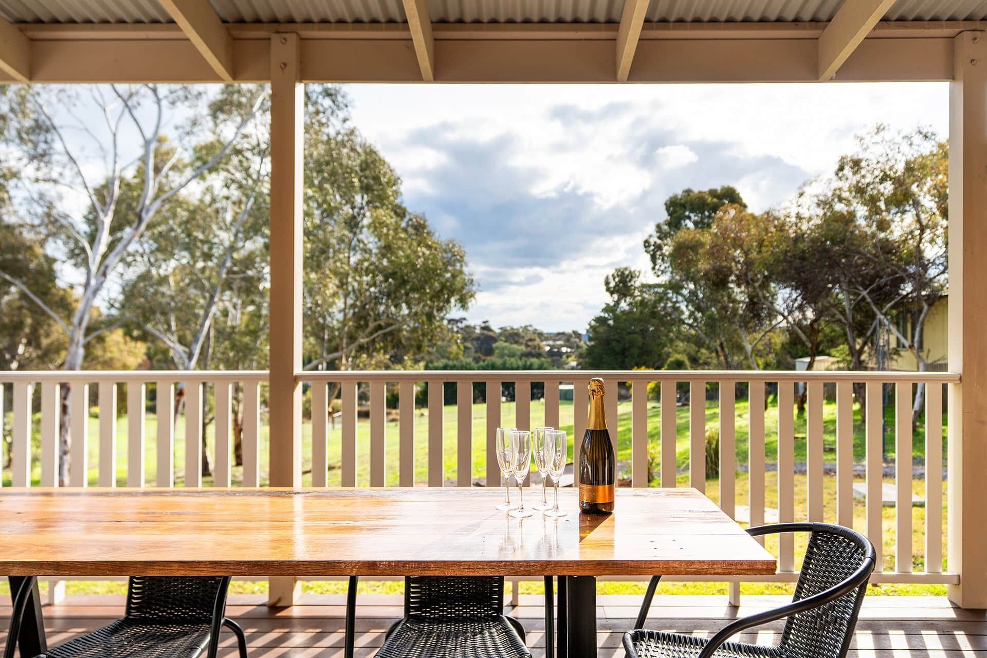 A wooden table with four chairs, a bottle of champagne, and three glasses on a covered patio with a view of a grassy field and trees.