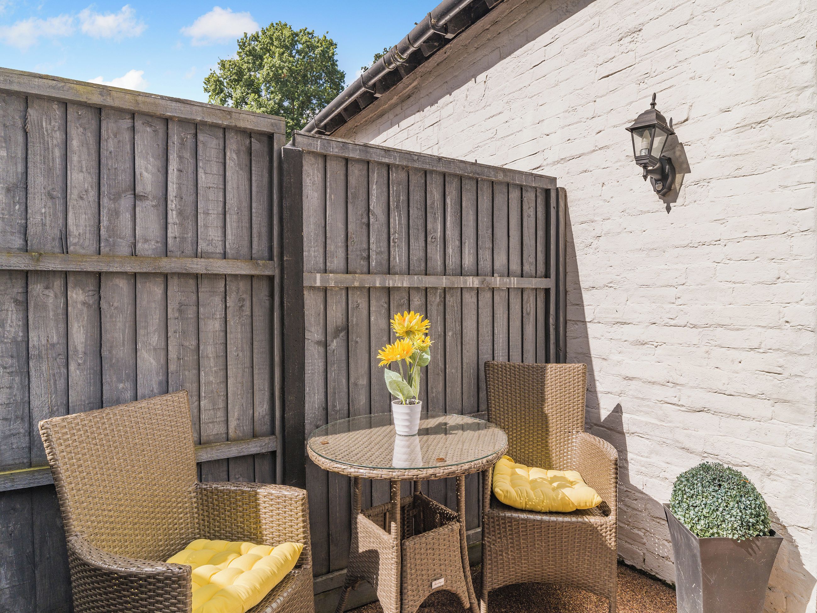 Eine Terrasse mit Korbstühlen, einem Glastisch und einer Vase mit Sonnenblumen.