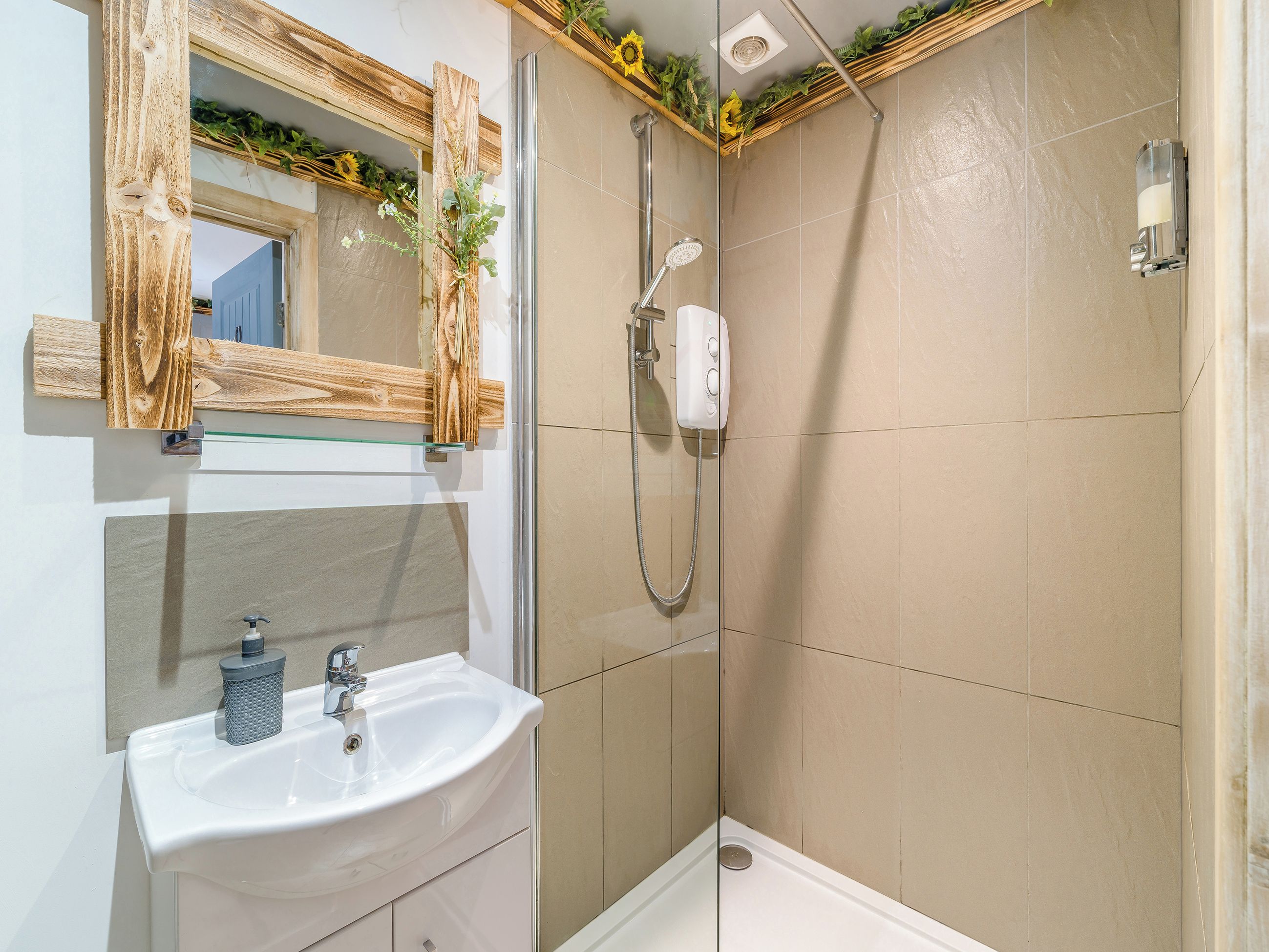 Un baño con un lavamanos blanco, un espejo con marco de madera, una ducha con cabezal de ducha manual y paredes con azulejos color beige.