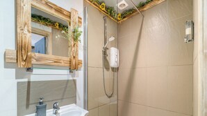Un baño con un lavamanos blanco, un espejo con marco de madera, una ducha con cabezal de ducha manual y paredes con azulejos color beige.
