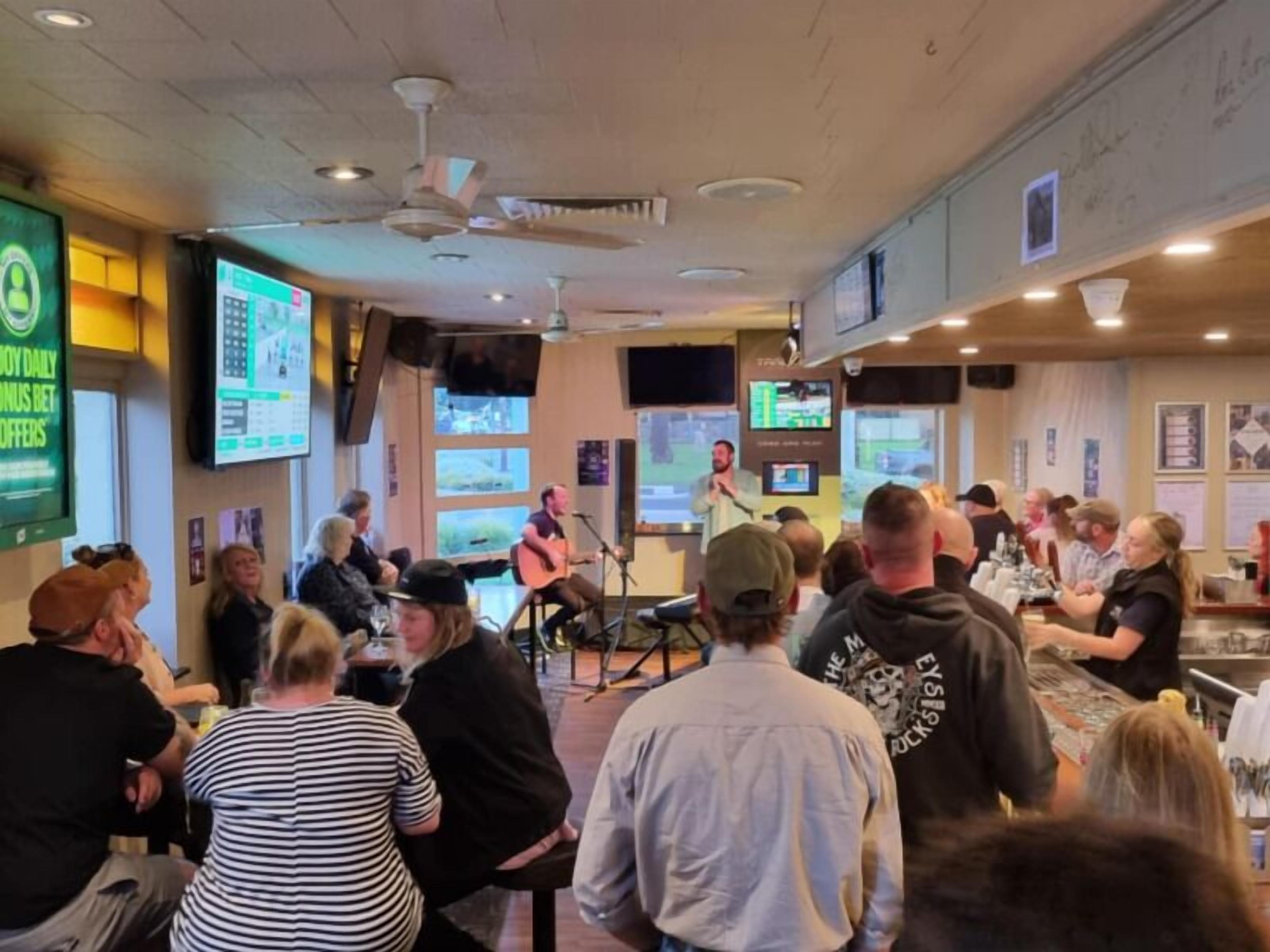A crowded indoor space with multiple screens displaying content, a stage with a performer, and people seated and standing around.