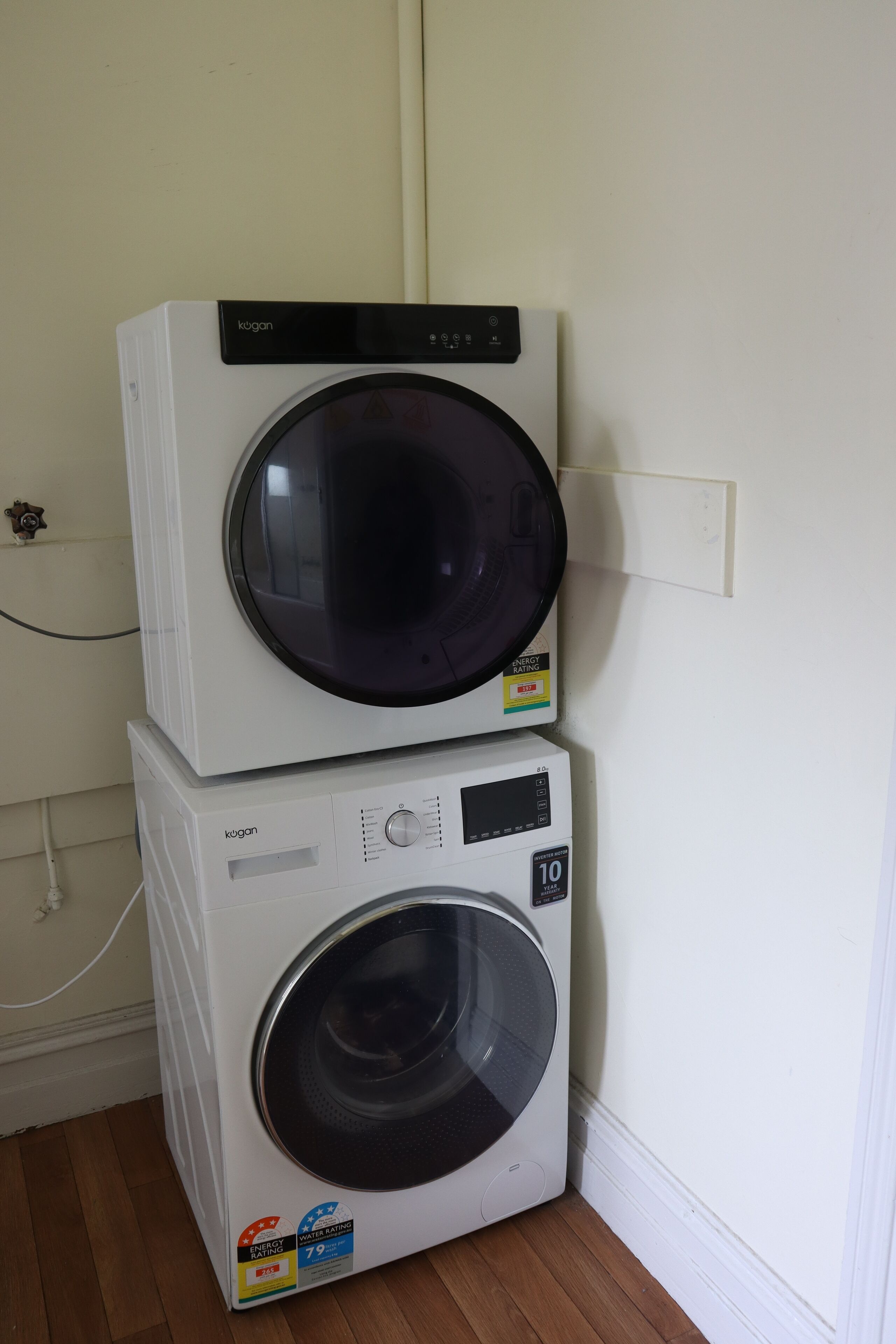 Two stacked white washing machines with a control panel and a digital display, placed against a wall with a power outlet.