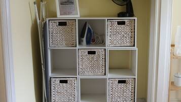 A corner of a hotel room with a white shelving unit containing woven baskets and a framed picture.