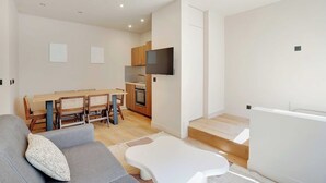 Un salon moderne avec un canapé gris, un pouf blanc et un coin repas comprenant des chaises et une table en bois.