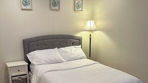 Ein ordentlich bezogenes Bett mit weißen Leinen, ein grauer möbelbezogener Kopfteil und ein Nachttisch mit Lampe.