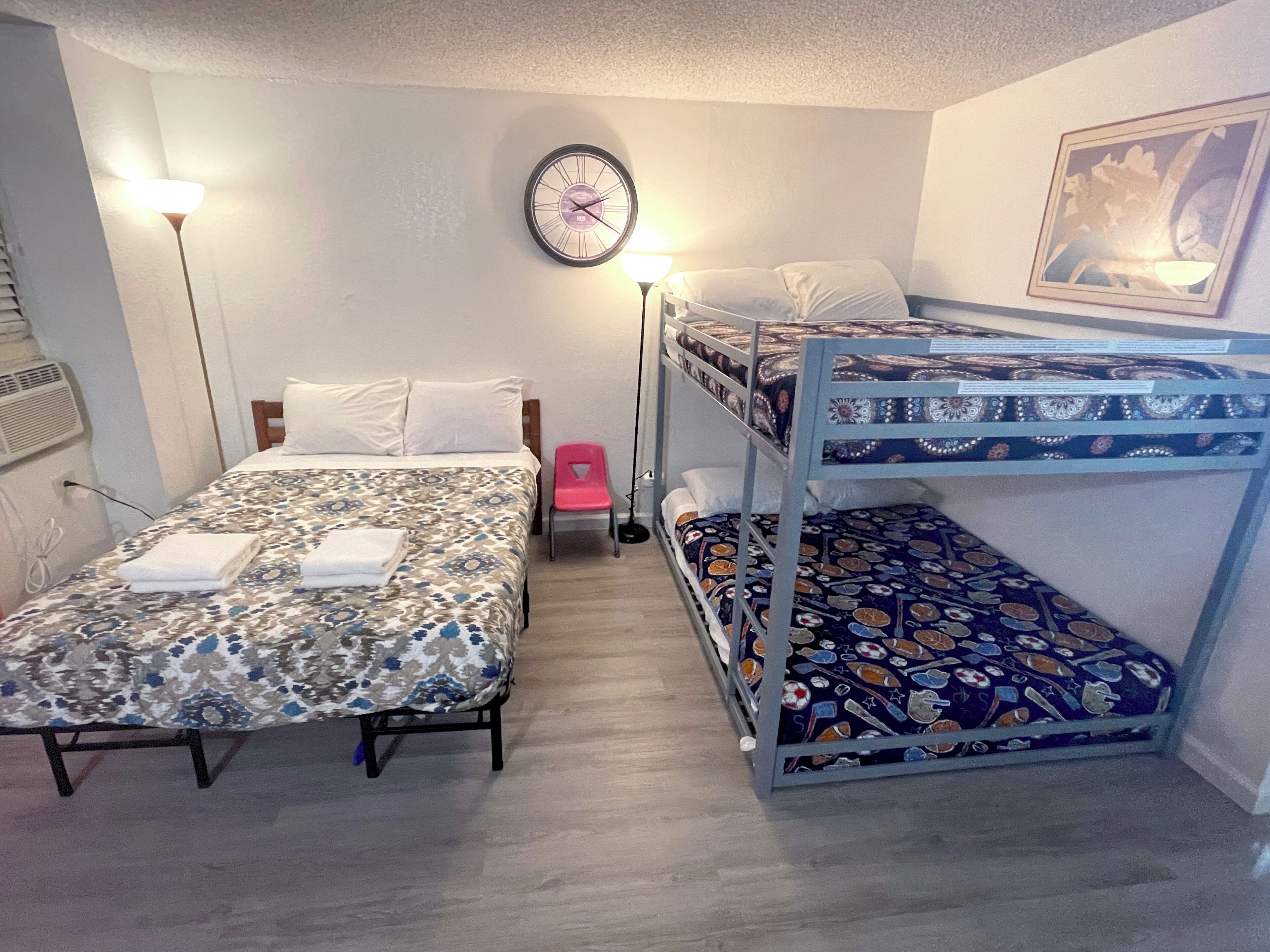 Een kamer met twee eenpersoonsbedden, een stapelbed, een wandklok, een kleine stoel en een ingelijst schilderij.