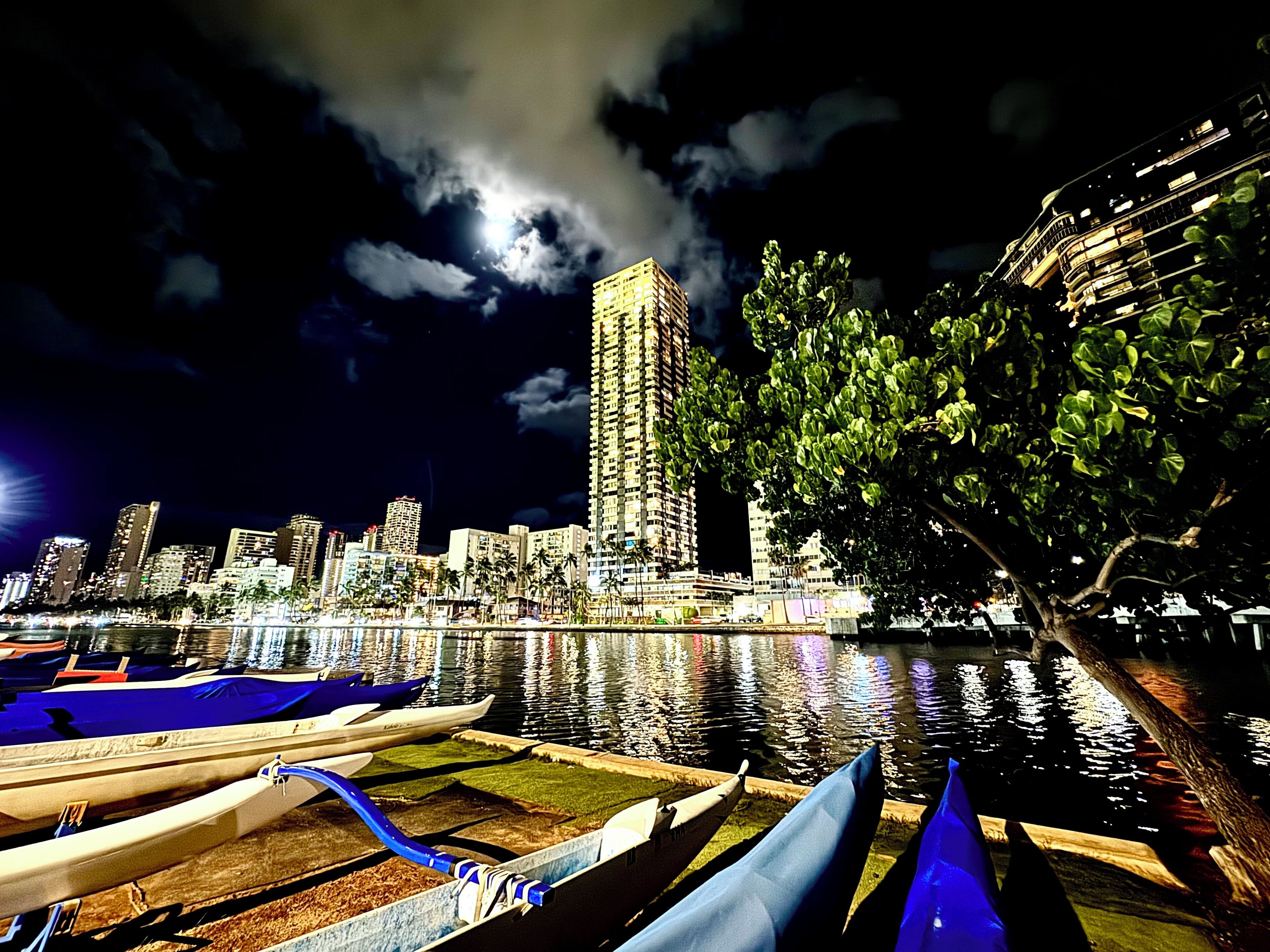 A nighttime cityscape with a prominent high-rise building, a calm water body reflecting lights, and a tree-lined area.