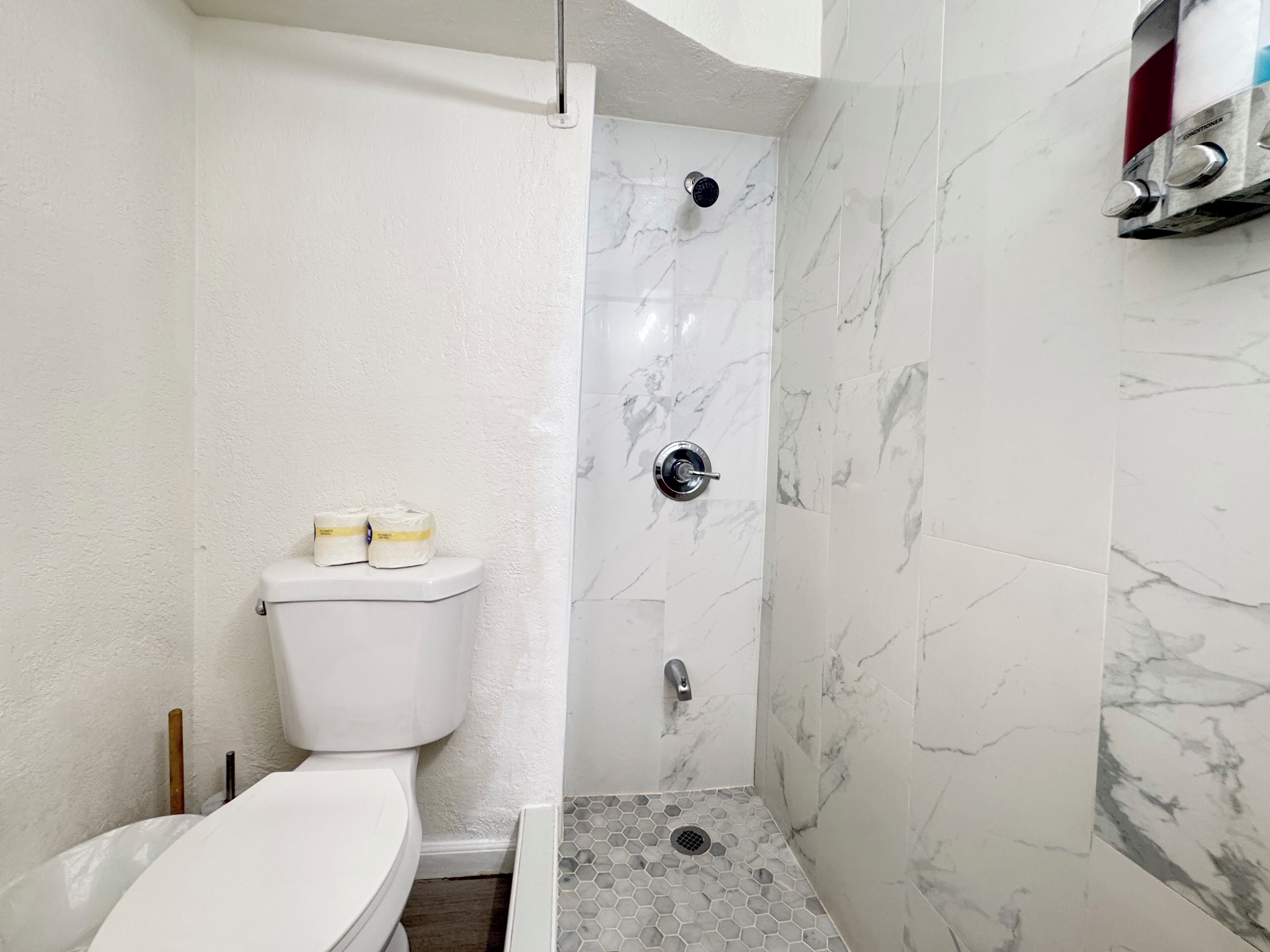 Et badeværelse med toilet, brusebad med marmorvæg og gulv med sekskantede fliser.