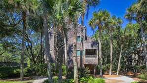 A rustic wooden house surrounded by tall palm trees and dense foliage. - Ocean Views, Serene Back Deck, Easy Walk to Beach. Beautiful Seaside, Dog-Friendly Villa Awaits You! (Kiawah Island)