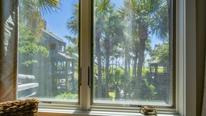A window with a view of a house and palm trees. - Ocean Views, Serene Back Deck, Easy Walk to Beach. Beautiful Seaside, Dog-Friendly Villa Awaits You! (Kiawah Island)