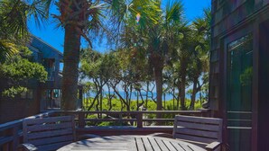 A wooden deck with a round table and benches, overlooking a scenic view of palm trees and a body of water. - Ocean Views, Serene Back Deck, Easy Walk to Beach. Beautiful Seaside, Dog-Friendly Villa Awaits You! (Kiawah Island)