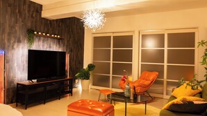 Un salon moderne doté d’un téléviseur à écran plat, d’un pouf en cuir, d’un fauteuil orange et d’une grande fenêtre avec des panneaux de verre dépoli.
