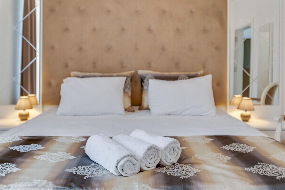 A neatly made bed with white linens and rolled towels, a tufted headboard, and bedside lamps.