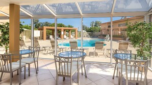 Un espacio al aire libre techado con mesas y sillas, con vista a una piscina y un edificio con pérgola.