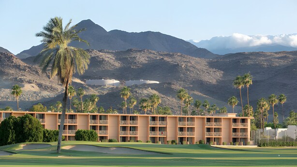 Campo de golf con un hotel al fondo, rodeado de montañas y palmeras.