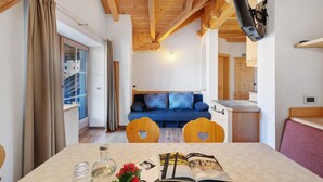 A room with a wooden ceiling, a blue sofa, a table with a magazine and a bottle, and a TV mounted on the wall. - Apartment 'Rît' with Mountain View, Shared Terrace and Wi-Fi (La Valle)