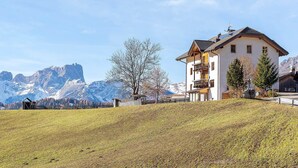 A two-story building with a sloped roof, a fenced yard, and mountains in the background. - Apartment 'Rît' with Mountain View, Shared Terrace and Wi-Fi (La Valle)