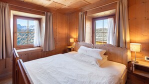 A wooden room with a bed, two windows with curtains, a nightstand with a lamp, and a view of mountains. - Apartment 'Ciablun Sas Dla Crusc' with Mountain View, Shared Terrace and Wi-Fi (La Valle)