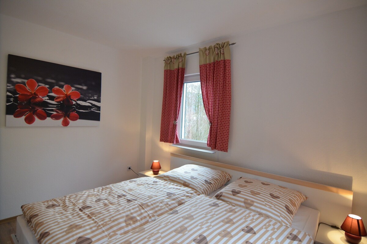A bedroom with a bed, two bedside lamps, a window with red curtains, and a wall-mounted painting.