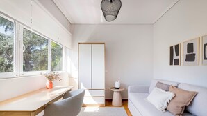 A modern living room with a desk, a grey chair, a white sofa with cushions, a small round table with a lamp, and a large window with a view of trees. - Family Escape in Chalandri -Parnassidos 10 (Chalandri)